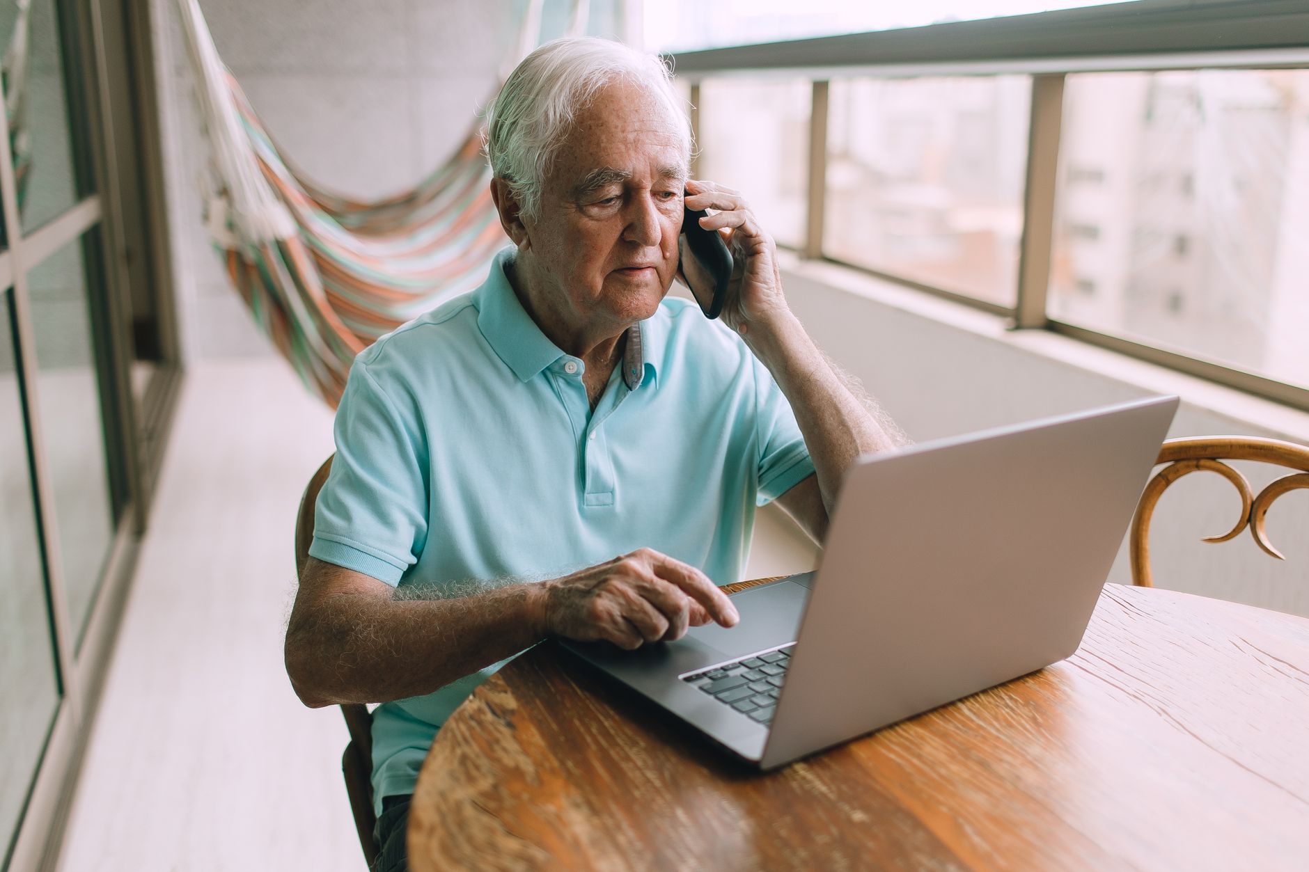 Older man on a phone call while using a laptop at a wooden table by a window