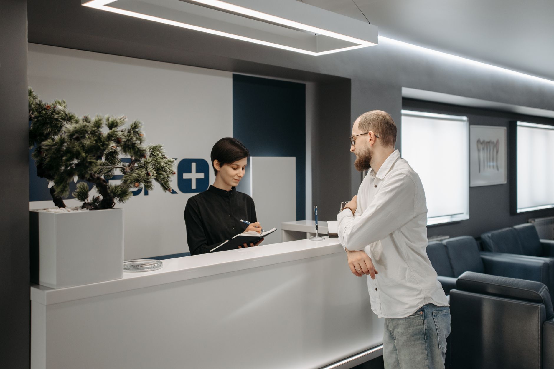 A man in a white shirt talks with a staff member who takes notes at an office reception counter, with a row of waiting-room chairs behind him