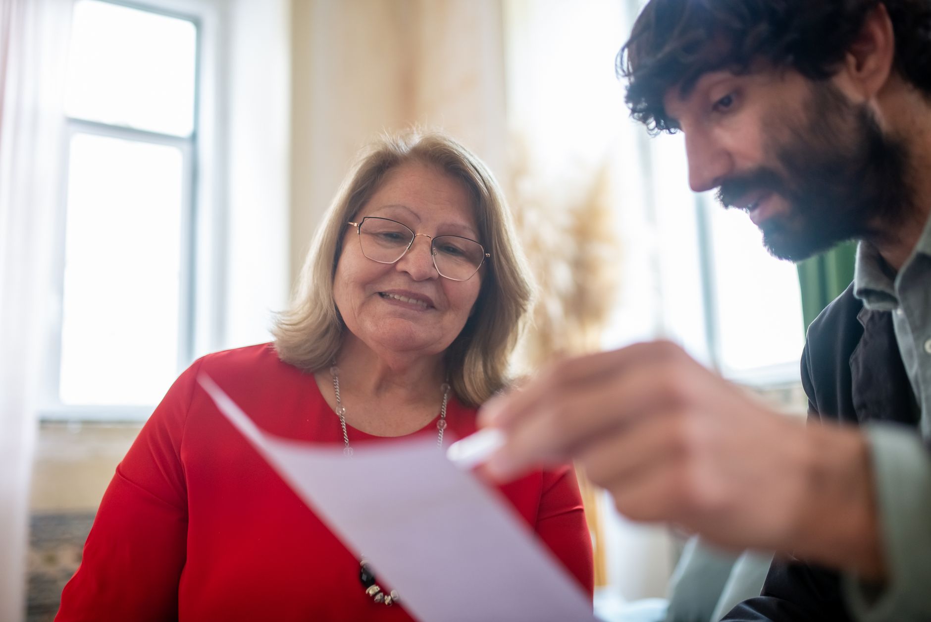 An older woman in glasses smiles as a younger man beside her holds up a sheet of paper they are reading together