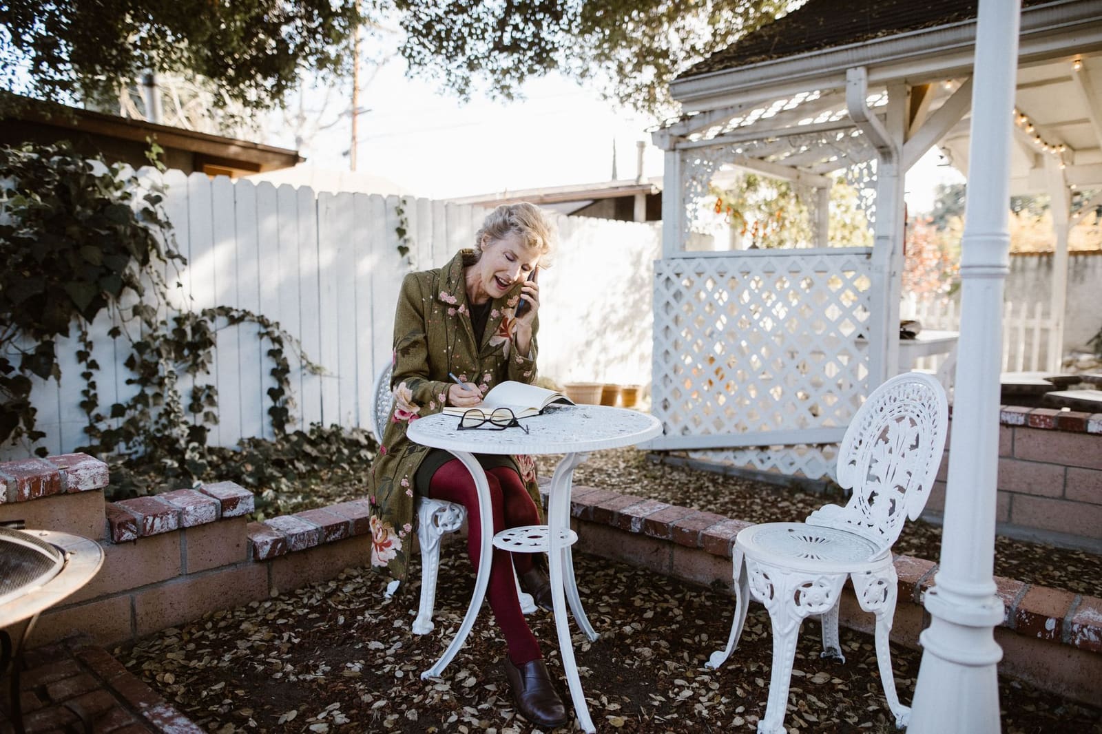An older woman at a garden table talks on her phone and writes notes in a notebook, her reading glasses on the table