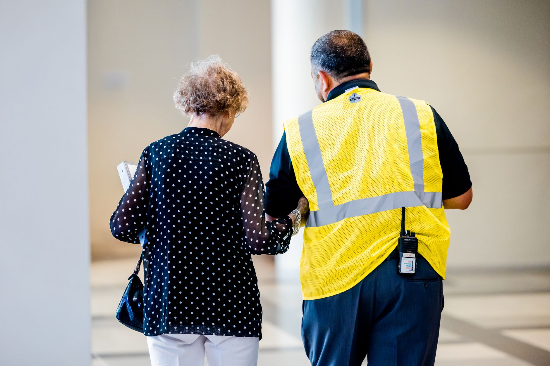 A security officer in a yellow vest walks beside an older woman carrying a folder through a bright building hallway