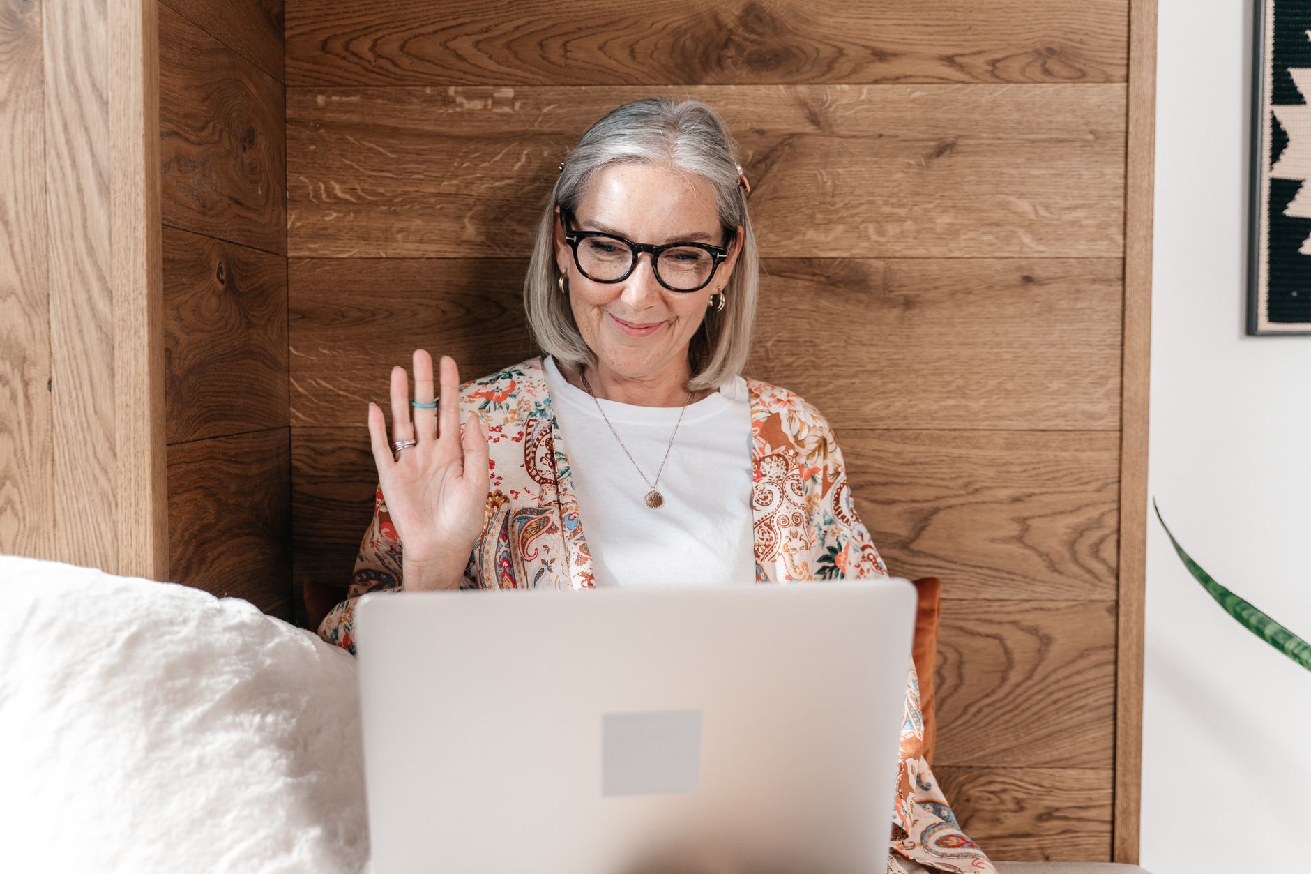 Older woman with glasses waving at a laptop screen during a video call at home