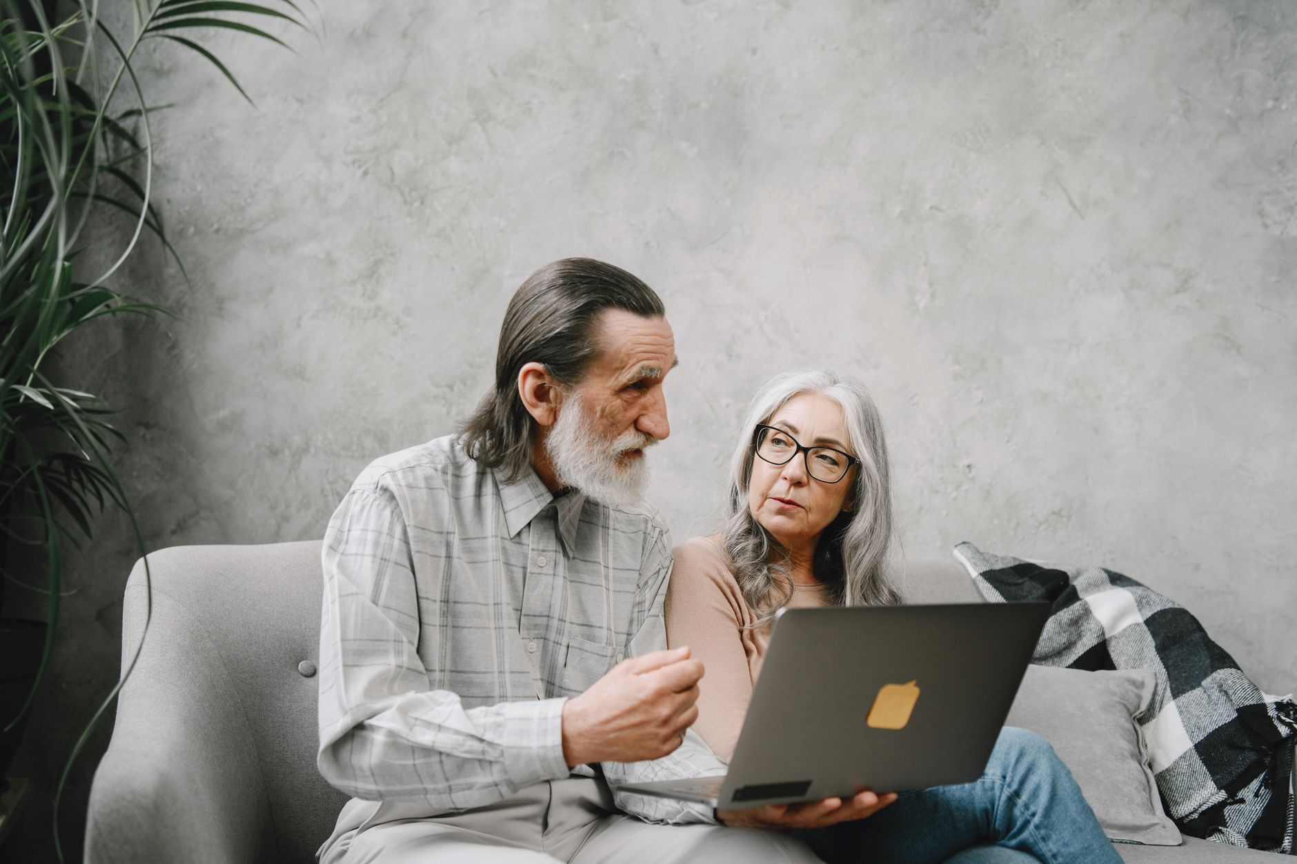 An older man and woman sit together on a gray sofa, looking something up on a laptop